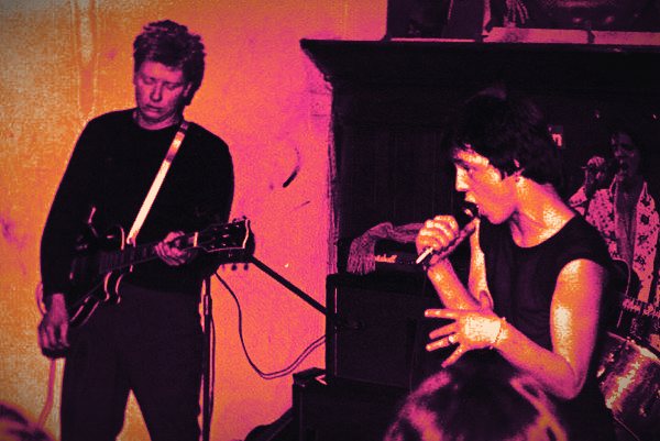 Plastix on stage: a guitarist on the left and a singer holding a microphone on the right, graded in orange and magenta